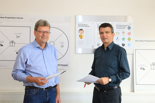 Zwei Männer, entspannt in Richtung des Betrachters schauend, im Hintergrund Visualisierungen Mindmaps