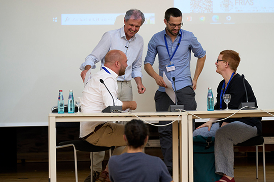 Vier Personen auf der Bühne. Zwei Personen sitzen und sind den zwei hinter ihnen stehenden Personen zugewandt. Alle lachen und machen einen entspannten Eindruck.