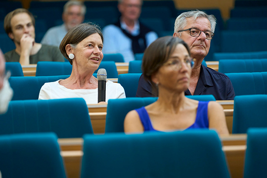 Menschen sitzen in Stuhlreihen. Eine Frau aus dem Publikum hält ein Mikro und spricht.